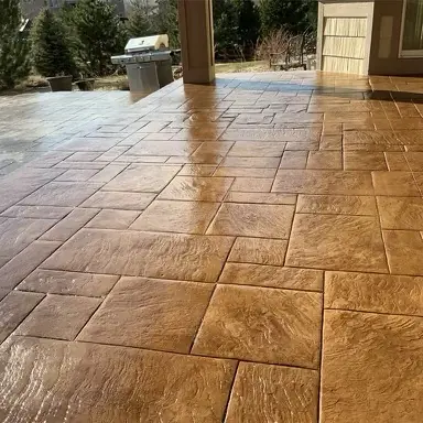 A stamped concrete patio in golden tones with pine trees and outdoor kitchen.